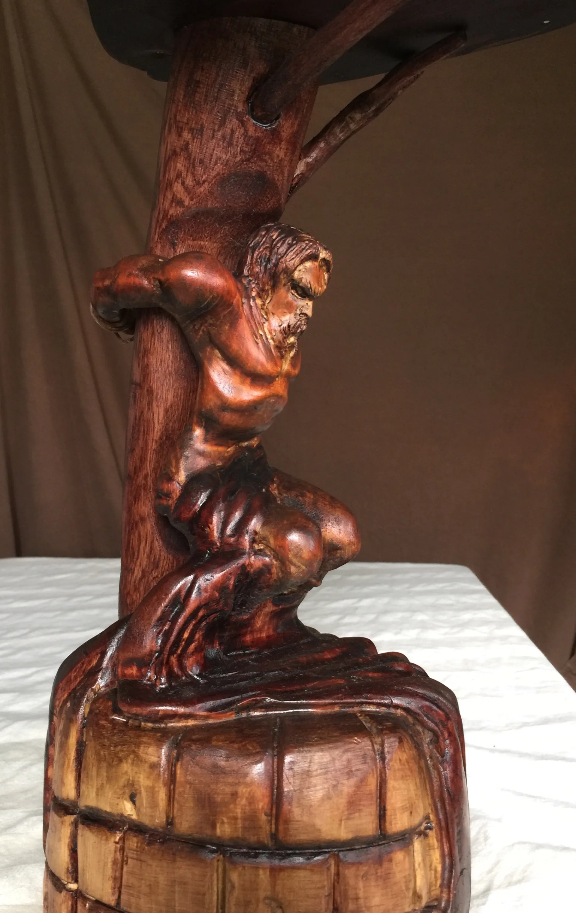 A carved wooden sculpture of a muscular crouching figure gripping a wooden post, mounted on a barrel-shaped base.