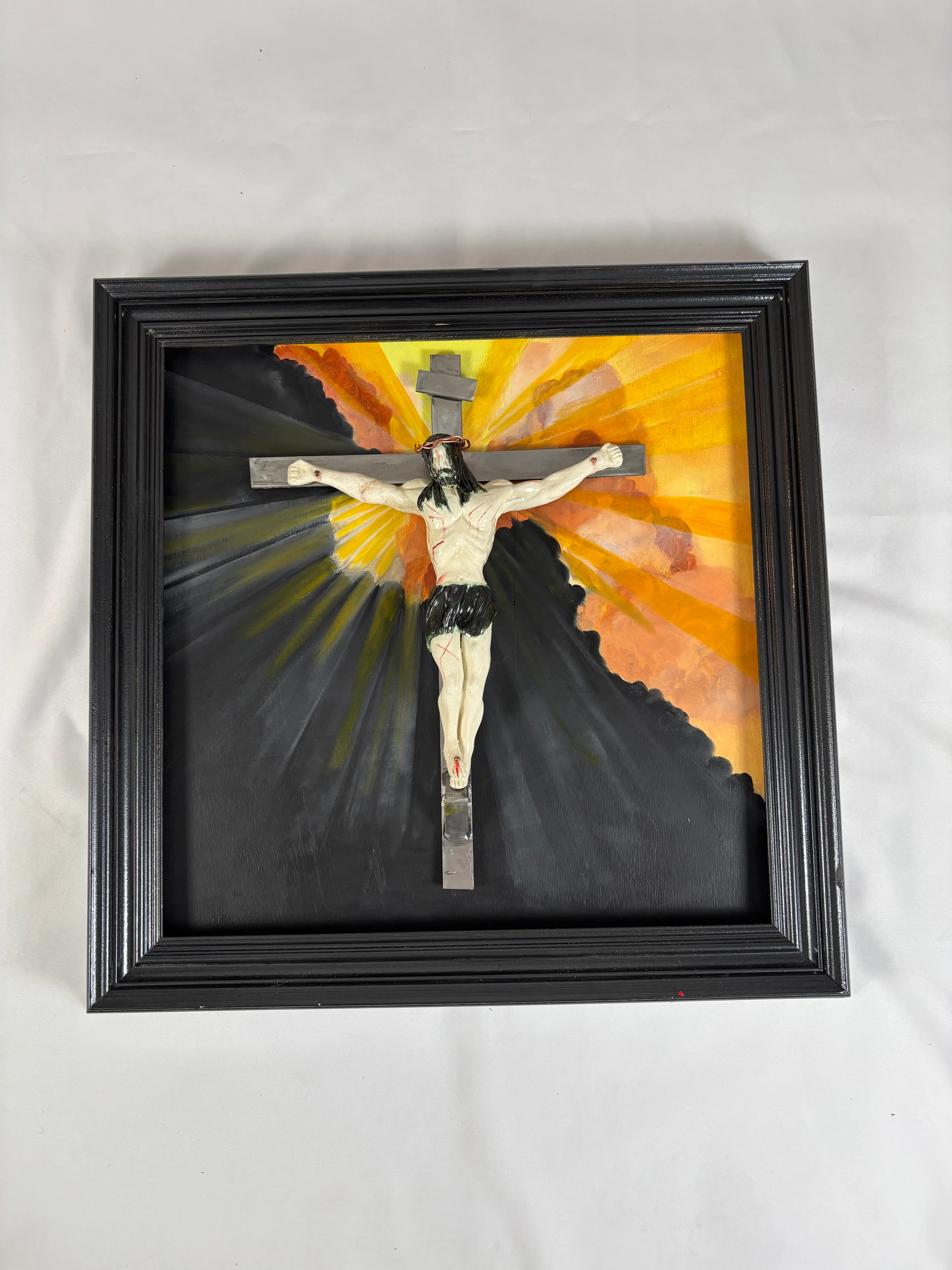 A framed mixed-media artwork of a crucifix with a sculpted figure against painted rays of light.