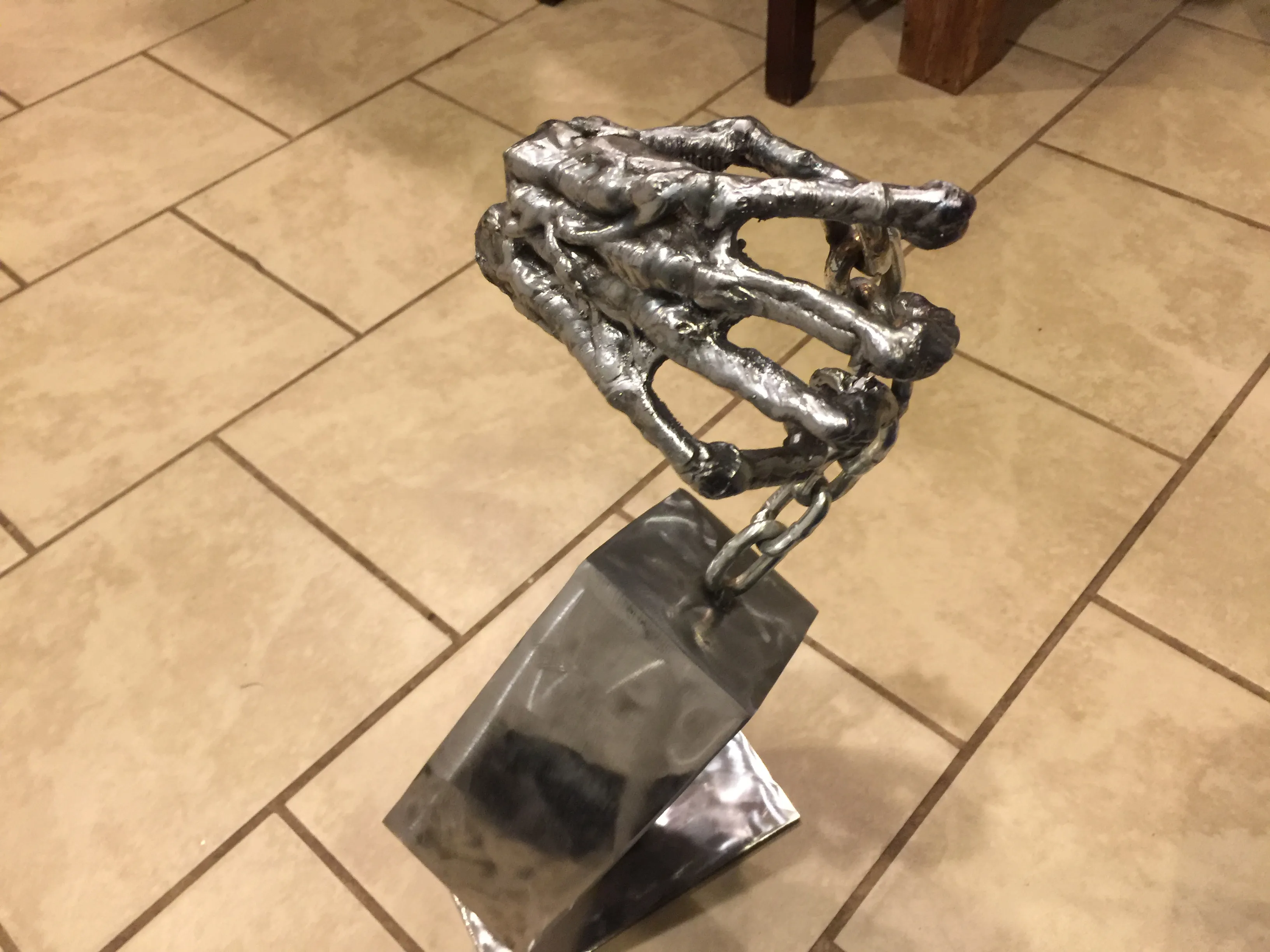 A welded metal skeletal hand sculpture connected by a chain to an angled metal base.