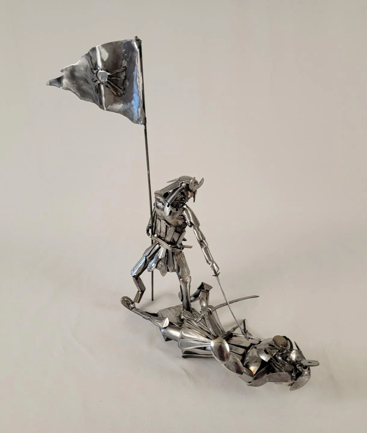This piece brings together intricate metalwork and dramatic storytelling—featuring a horned samurai standing over a fallen enemy, all assembled from welded spoons, forks, and repurposed flatware.